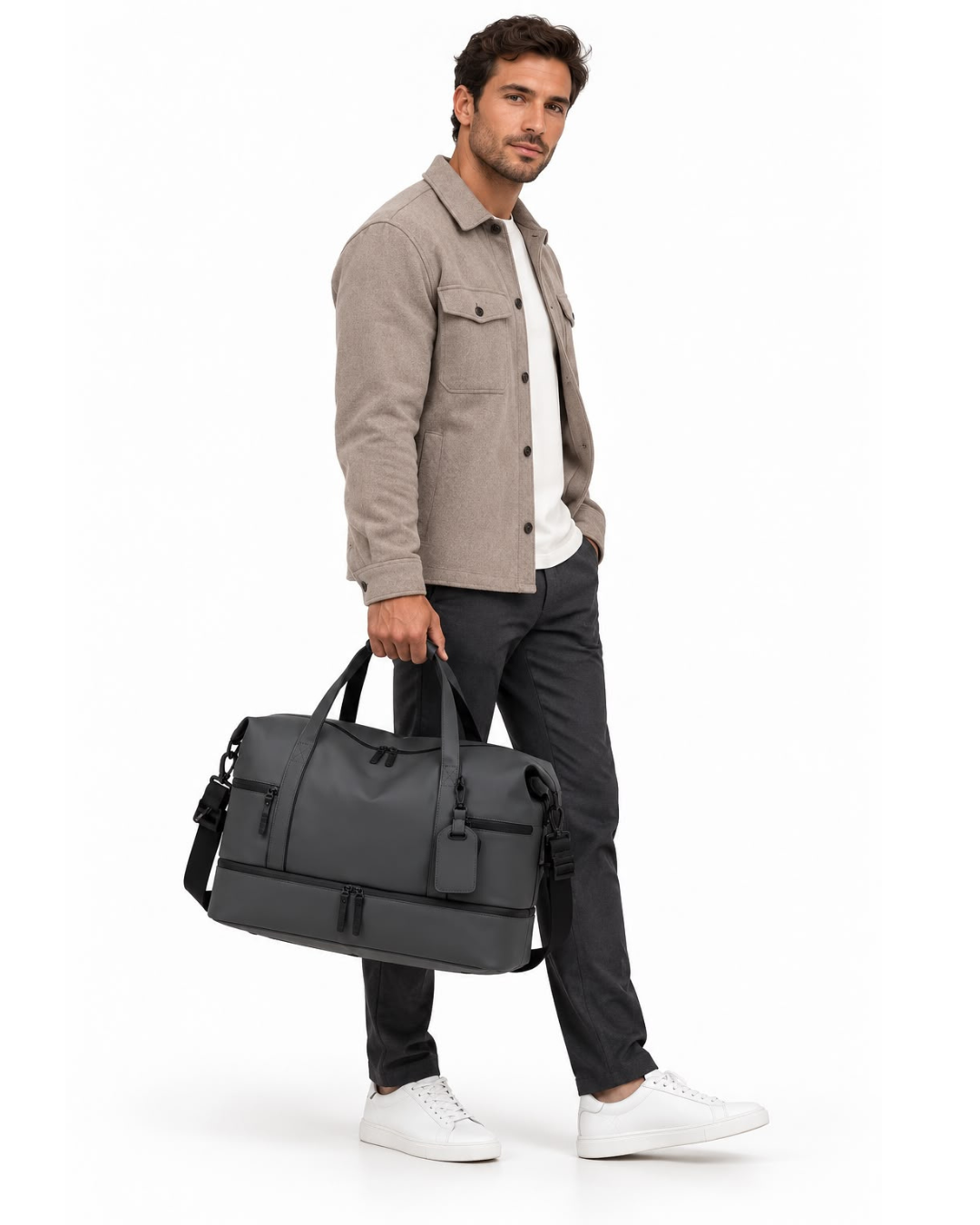 Man carrying the dark grey AVREN men's travel duffle bag with top handles, an adjustable shoulder strap, multiple zippered pockets and a dedicated shoe compartment, styled in smart casual clothing against a white studio background.